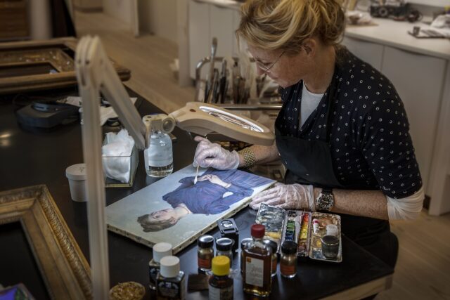 A conservator carefully cleaning a portrait painting under a magnifying lamp.