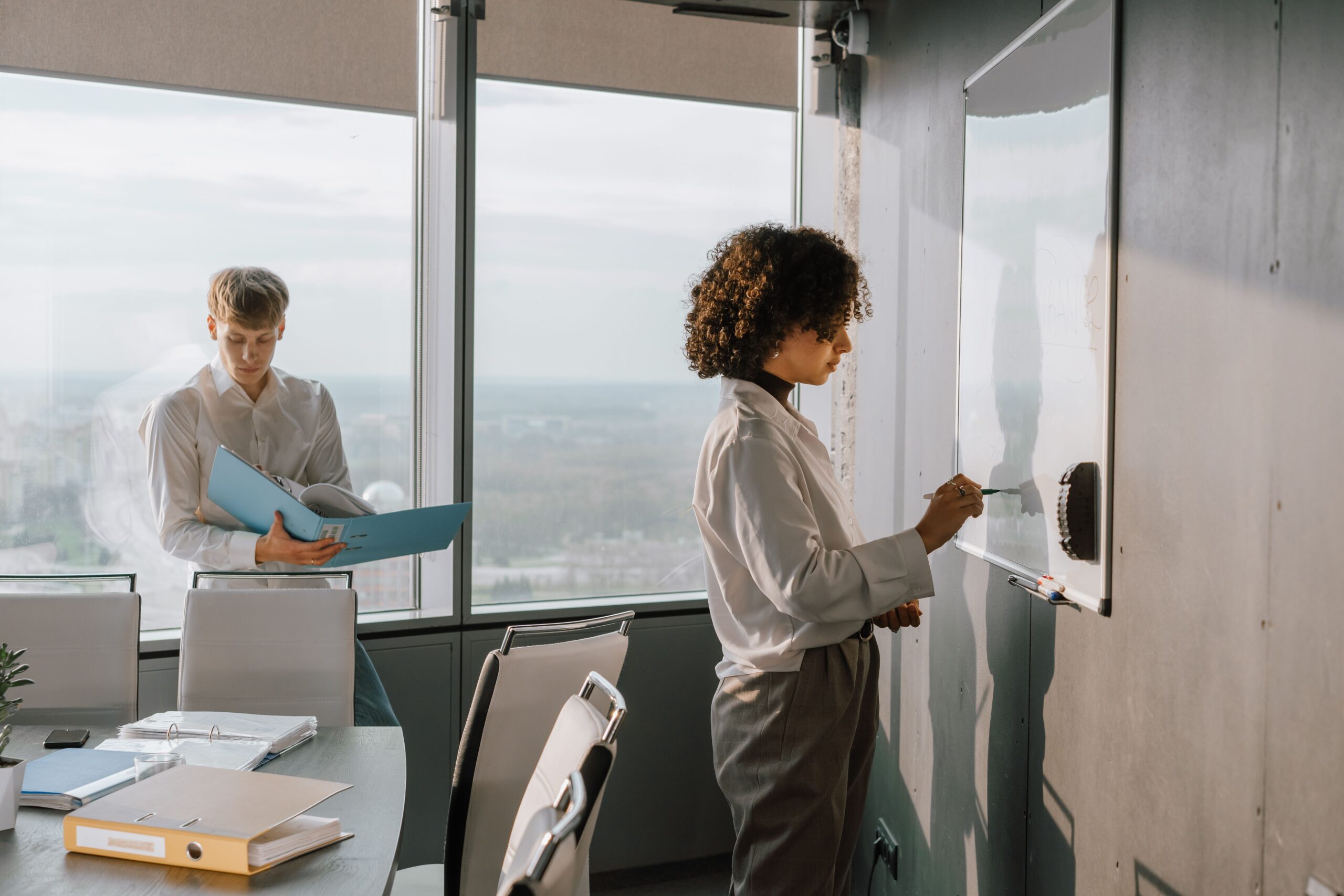 Two colleagues planning together in an office, with one writing on a whiteboard while the other reviews project documents.